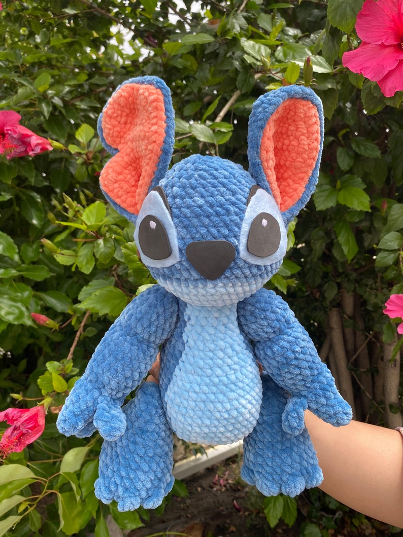 May include: A handmade blue crochet plush toy with large ears, featuring orange inner ears, and black eyes. The plush has a light blue belly and a black nose. The toy is held in front of a green leafy background with pink flowers.