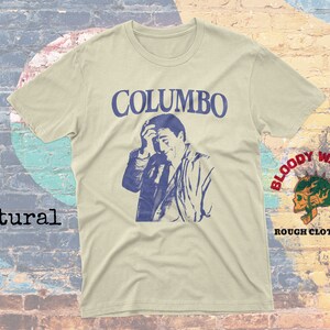 May include: A beige t-shirt with a blue and white graphic print of a man with the text "COLUMBO" above him. The shirt has a small patch on the right sleeve with the text "BLOODY WASABI" and "ROUGH CLOTHING" below it.