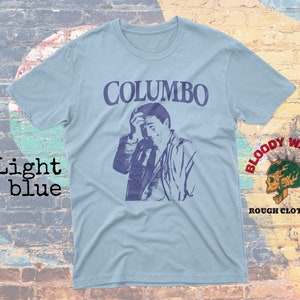 May include: Light blue t-shirt with a graphic print of actor Peter Falk as Columbo, a fictional detective. The text "COLUMBO" is printed in black above the image. The t-shirt is from the brand "Bloody Wasabi Rough Clothing."