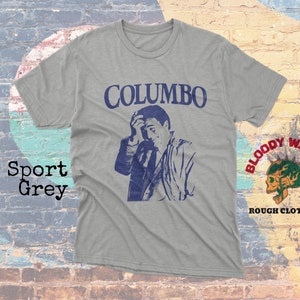 May include: A gray t-shirt with a blue and white image of actor Peter Falk as Columbo, with the text "COLUMBO" above the image. The shirt also features a small logo in the bottom right corner that says "BLOODY WASABI ROUGH CLOTHING".