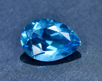 May include: A pear-shaped blue topaz gemstone with a faceted cut. The gemstone is set against a dark blue background.
