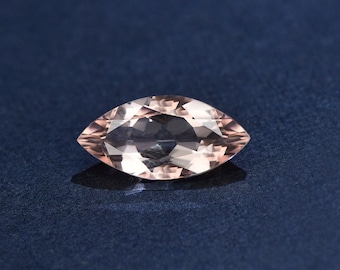 Puede incluir: Una piedra preciosa de color rosa claro tallada en forma de marquesa con facetas. La piedra preciosa está sobre un fondo azul oscuro.