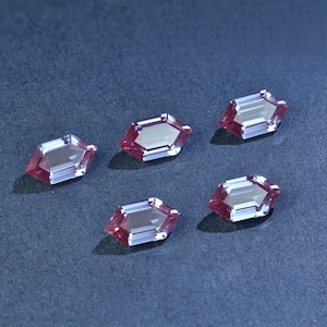 May include: Six hexagonal-shaped gemstones with a reddish-purple hue. The gemstones are arranged in a row on a dark gray surface.