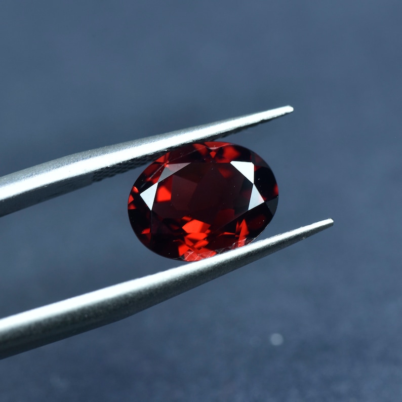 May include: A deep red oval gemstone held by tweezers against a blue background.