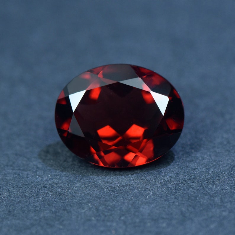 May include: A deep red gemstone with a faceted oval shape. The gemstone is set against a dark blue background.