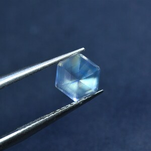 May include: A clear, hexagonal gemstone with a rainbow sheen, held by tweezers against a blue background.