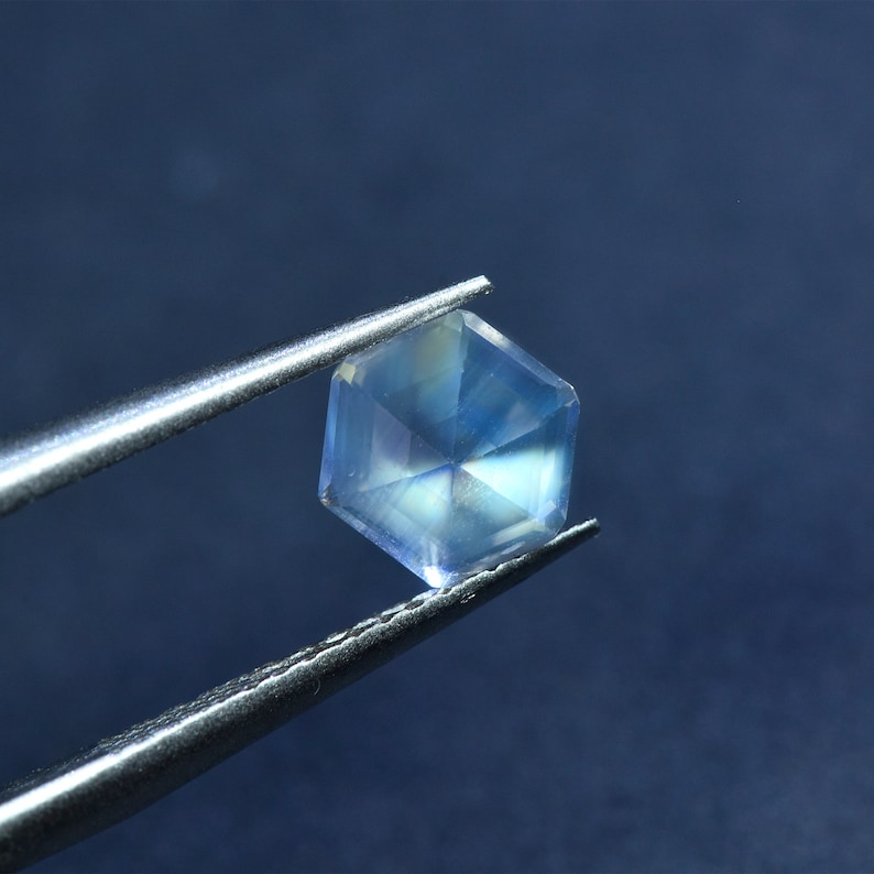 May include: A clear, hexagonal gemstone with a rainbow sheen, held by tweezers against a blue background.
