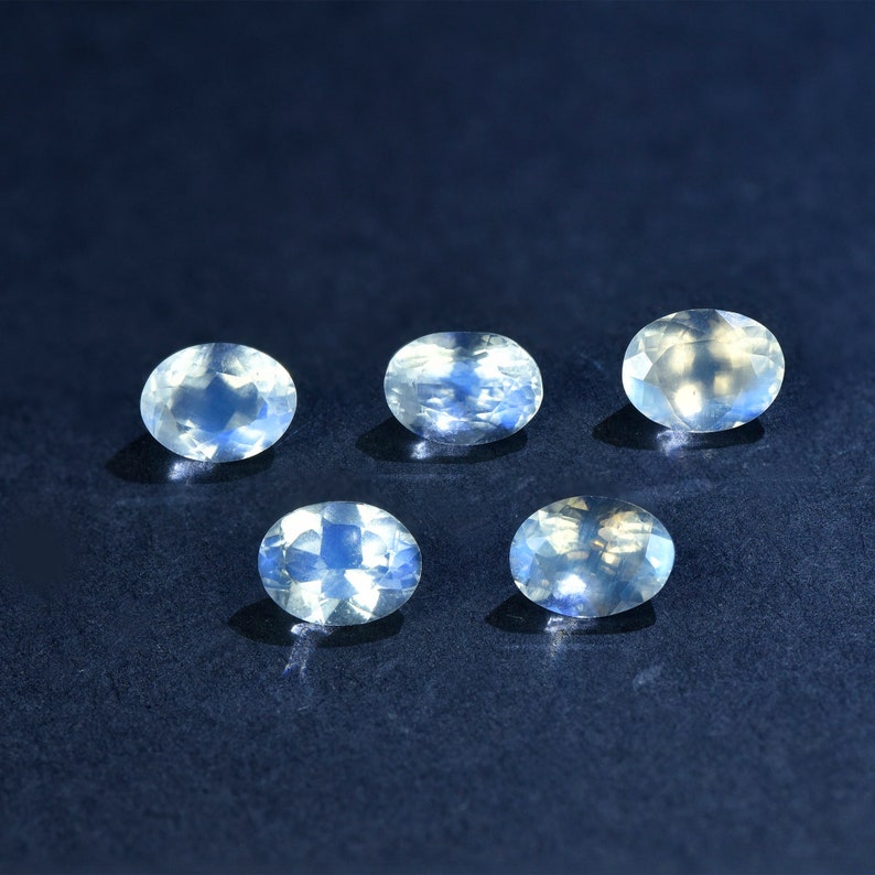 May include: Six oval-shaped moonstone gemstones with a milky white color and iridescent blue flashes. The stones are arranged in two rows of three on a dark blue background.