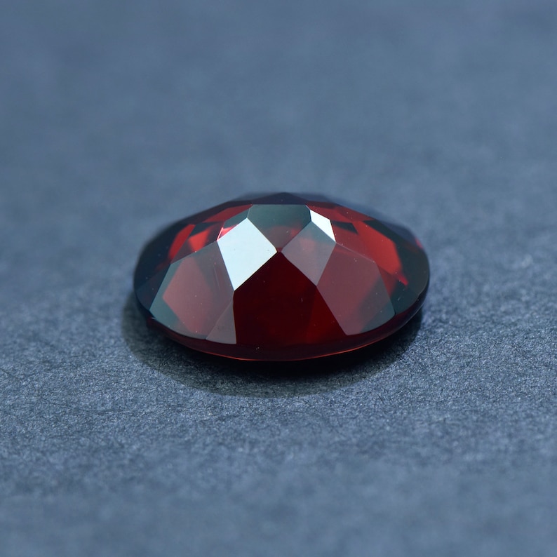 May include: A deep red gemstone with a faceted cut. The gemstone is oval shaped and has a high polish.