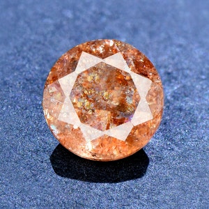 May include: A round, faceted gemstone with a reddish-brown colour and a starburst pattern. The gemstone is set against a blue background.