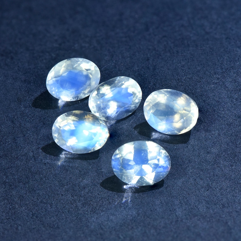 May include: Five oval-shaped moonstone gemstones with a milky white color and iridescent blue flashes.