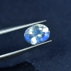 May include: A clear oval-shaped gemstone with a blue iridescent sheen, held by tweezers against a dark blue background.