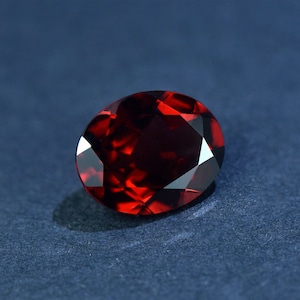 May include: A deep red gemstone with a faceted oval shape. The gemstone is set against a dark blue background.