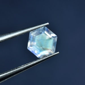 May include: A clear, iridescent gemstone with a hexagonal shape, held by tweezers against a blue background.