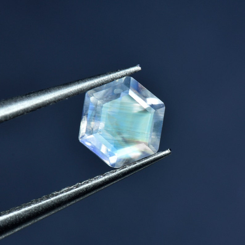 May include: A clear, iridescent gemstone with a hexagonal shape, held by tweezers against a blue background.