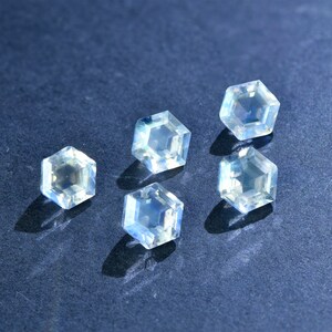 May include: Five iridescent, hexagonal, faceted gemstones on a dark blue background. The gemstones are clear with a slight blue hue.