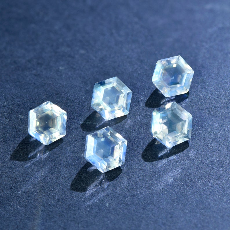 May include: Five iridescent, hexagonal, faceted gemstones on a dark blue background. The gemstones are clear with a slight blue hue.