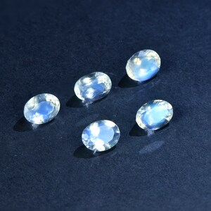 May include: Five oval-shaped moonstone gemstones with a milky white color and iridescent blue and green flashes. The stones are arranged in a semi-circle on a dark blue background.
