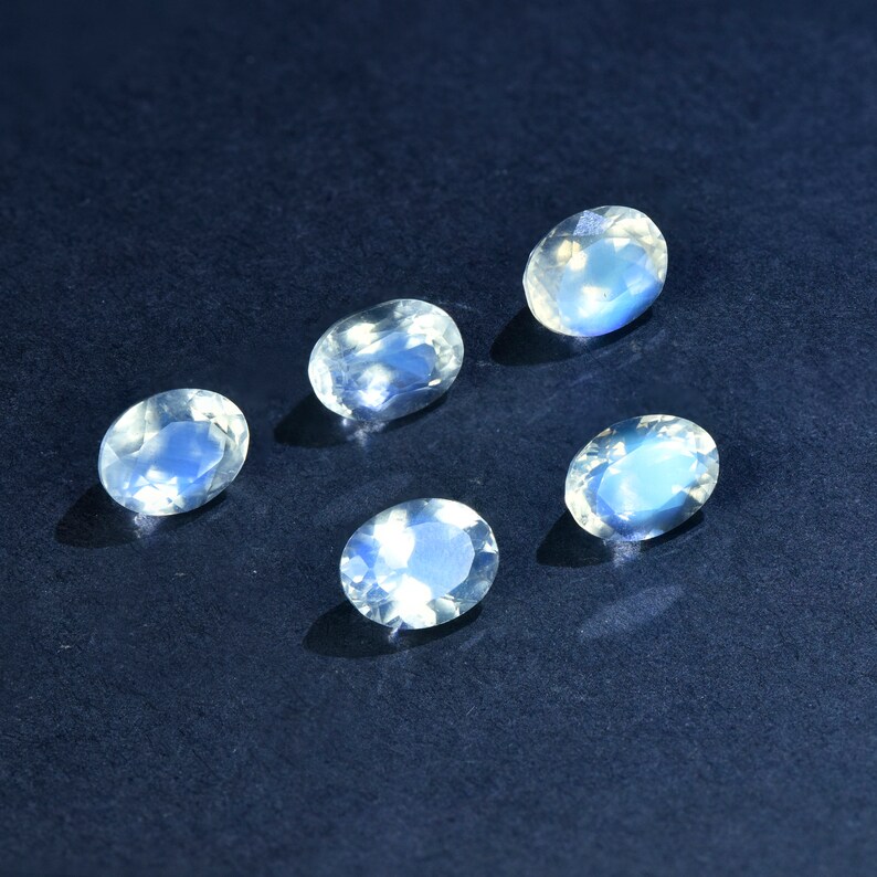 May include: Five oval-shaped moonstone gemstones with a milky white color and iridescent blue and green flashes. The stones are arranged in a semi-circle on a dark blue background.