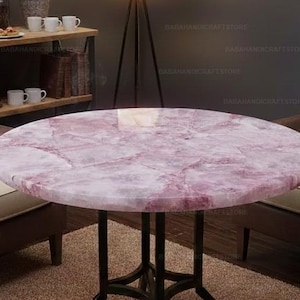Puede incluir: Una mesa redonda con una tapa de mármol rosa y blanco y patas de metal negro. La mesa está rodeada de sillones marrones con cojines color crema, creando una zona de estar acogedora. El diseño de la mesa añade un toque de elegancia al espacio.