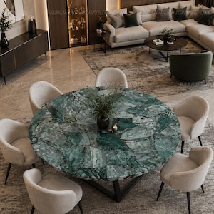 May include: A round dining table with a green marble mosaic top and a black metal base, surrounded by six light beige upholstered chairs with dark legs. A small green plant sits in the centre of the table on the polished stone surface.