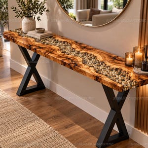 Könnte beinhalten: Ein langer Konsolentisch mit einer Tischplatte aus Naturholz, die in der Mitte eine flussartige Einlage aus glatten, bunten Kieselsteinen in klarem Harz aufweist. Der Tisch wird von zwei schwarzen Metallbeinen in X-Form getragen.
