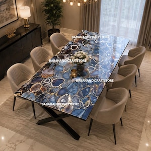 Puede incluir: Una mesa de comedor rectangular con una superficie de ágata azul pulida con variados patrones en azul oscuro, blanco y tonos dorados, sostenida por una base de metal negro en forma de X, rodeada de seis sillas beige tapizadas con patas oscuras.