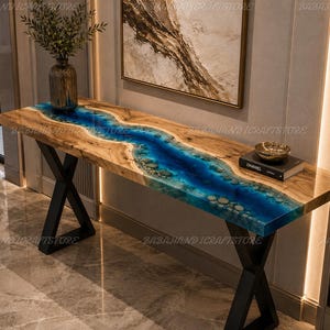 May include: A rectangular console table featuring a natural wood slab top with a central river-like inlay of translucent blue resin embedded with small stones. The table is supported by two black metal X-shaped legs and holds a vase and a book.