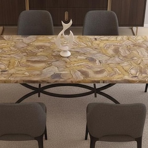 Puede incluir: Una mesa de comedor rectangular con una superficie estampada de color marrón claro, sostenida por una base de metal oscuro. Seis sillas grises tapizadas rodean la mesa. Una escultura decorativa blanca se encuentra sobre la mesa.