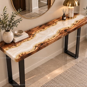 Può includere: Un tavolo console rettangolare con un piano in legno a bordo naturale e un inserto centrale in resina bianca. I bordi del legno sono arricchiti da dettagli in foglia d'oro. Il tavolo è sostenuto da due gambe in metallo nero a forma di U.