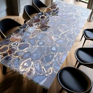May include: A rectangular dining table with a polished agate stone top in shades of blue, grey, and brown. The table is surrounded by six black chairs with wooden legs. The table's surface has a unique, natural pattern.