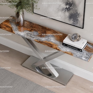 May include: A modern console table with a live edge wooden top featuring a river of silver resin. The table has a brushed metal base and is decorated with a vase, a geode, and books. The table is approximately 60 inches long.