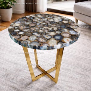 May include: A round side table featuring a mosaic top made of agate stones in shades of grey, white, and blue. The table has a gold-coloured metal base with an X-shaped design. The table is approximately 61 cm in diameter.
