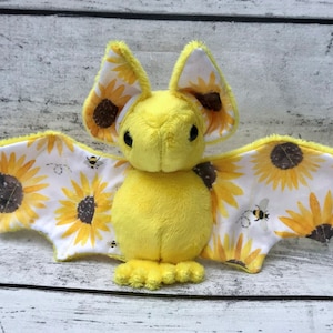 May include: A yellow plush bat with sunflower and bee patterned wings. The bat has black eyes and a small, rounded nose.