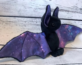 May include: A black plush bat with purple and blue galaxy-print wings. The bat has a soft, fuzzy texture and is a great addition to any collection.