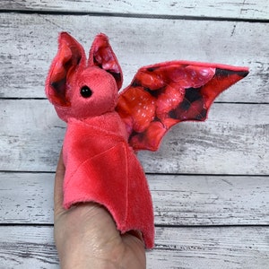 May include: A pink plush bat with a red and black patterned wing. The bat has black eyes and a pink body.