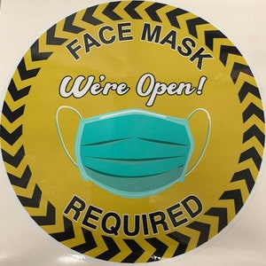 May include: Yellow round sign with black chevron border. The sign says "Face Mask Required" with a blue face mask graphic and the text "We're Open!" in a white cursive font.