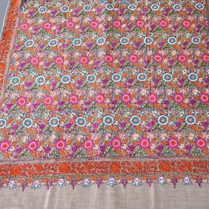 May include: An embroidered shawl featuring a floral design in pink, orange, blue, and purple. The border is a darker orange with detailed floral patterns. The background is a neutral beige colour.
