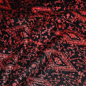 Könnte beinhalten: Schwarzer Stoff mit aufwendiger roter Blumenstickerei. Das detaillierte Muster zeigt zarte Ranken, Blätter und Blumenmotive, die ein reichhaltiges, strukturiertes Aussehen erzeugen. Der Stoff ist wahrscheinlich ein Schal oder Tuch.