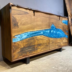 May include: A wooden cabinet with a striking blue resin river design. The cabinet is made of natural wood with a dark finish and features four doors. The blue resin flows across the front, creating a unique and artistic look. The cabinet sits on black legs.