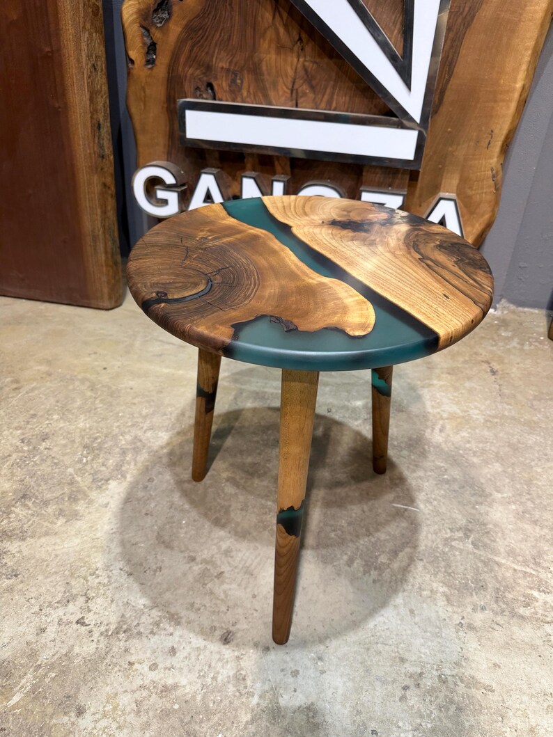 May include: A round wooden side table with a teal epoxy river design. The table has three wooden legs.
