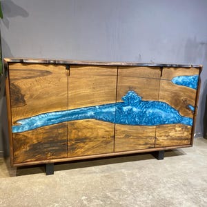 May include: A wooden cabinet with a striking blue resin river design. The cabinet features four doors and sits on black metal legs. The wood has a natural grain, and the blue resin adds a vibrant contrast.