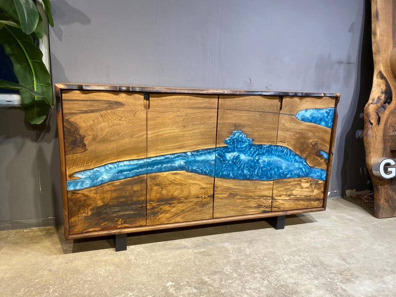 May include: A wooden cabinet with a striking blue resin river design. The cabinet features four doors and sits on black metal legs. The wood has a natural grain, and the blue resin adds a vibrant contrast.
