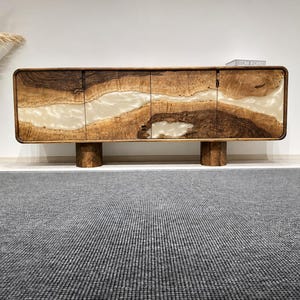Puede incluir: Un aparador rectangular de madera con diseño de borde natural y una banda horizontal central de resina blanca translúcida. El mueble tiene cuatro puertas y se apoya sobre dos patas cilíndricas de madera. Un libro descansa en la parte superior.