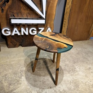 May include: A round wooden side table with a dark brown wood top and a teal blue epoxy river design. The table has three legs made of the same dark brown wood.