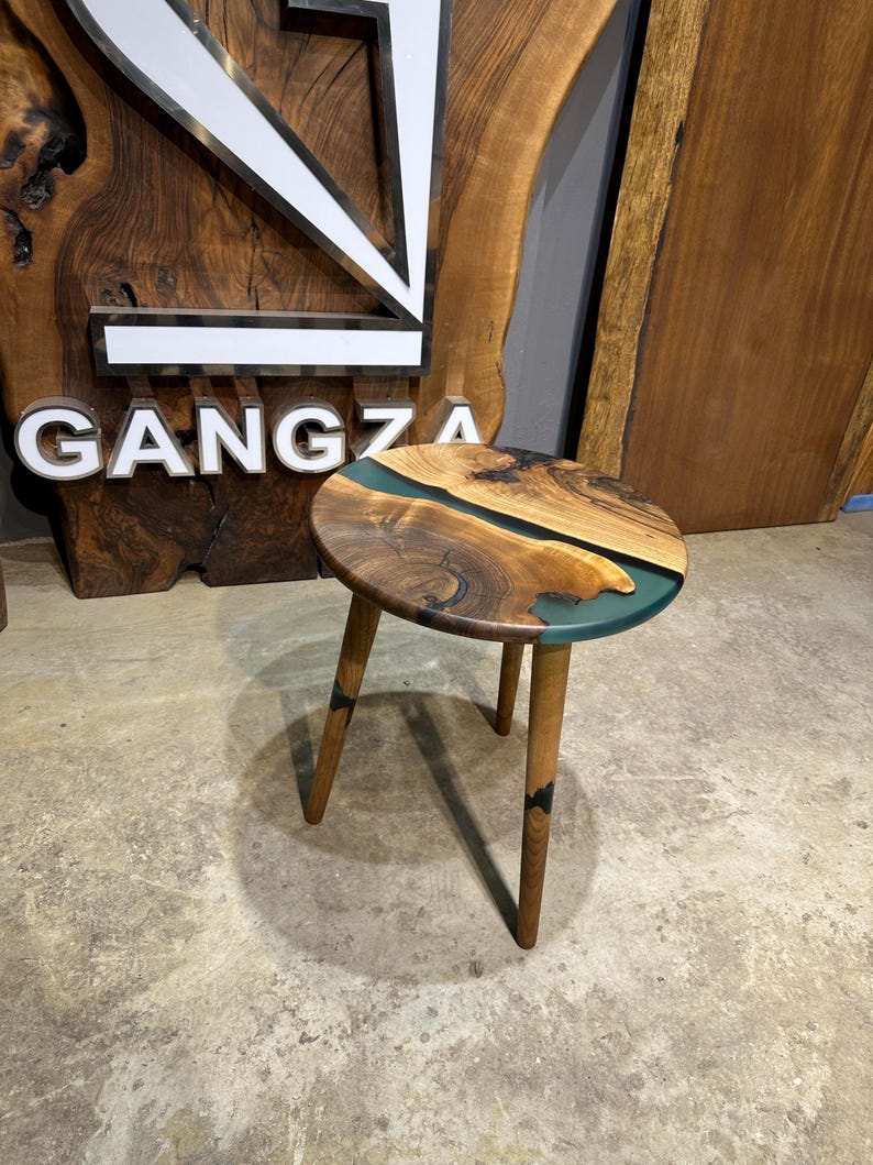 May include: A round wooden side table with a dark brown wood top and a teal blue epoxy river design. The table has three legs made of the same dark brown wood.