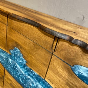 May include: A wooden cabinet with a natural edge and a river of blue epoxy resin. The cabinet is made of light brown wood with a dark brown edge. The blue resin flows through the wood, creating a unique and artistic design.