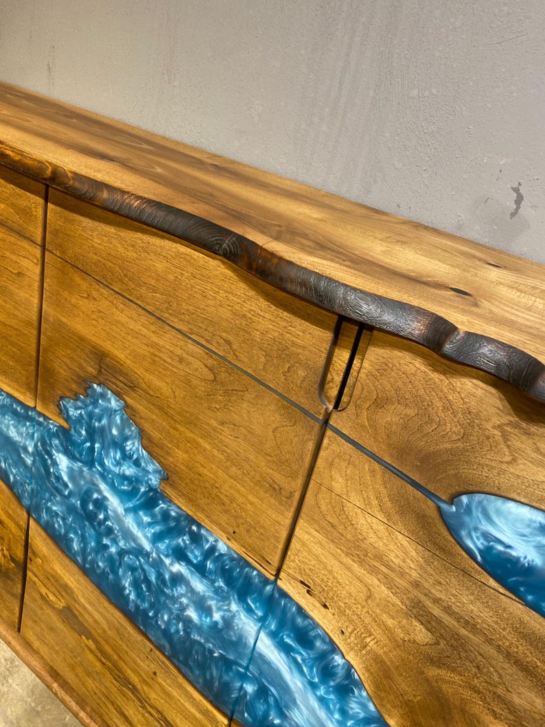 May include: A wooden cabinet with a natural edge and a river of blue epoxy resin. The cabinet is made of light brown wood with a dark brown edge. The blue resin flows through the wood, creating a unique and artistic design.