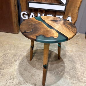 May include: A round wooden side table with a teal epoxy river design. The table has three wooden legs.