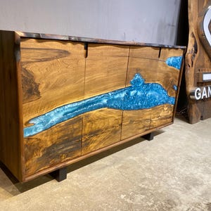 May include: A wooden cabinet with four doors and a striking blue resin river design. The cabinet is made of natural wood with a dark finish and sits on black legs. The word "GANGZA" is visible on a wooden sign.